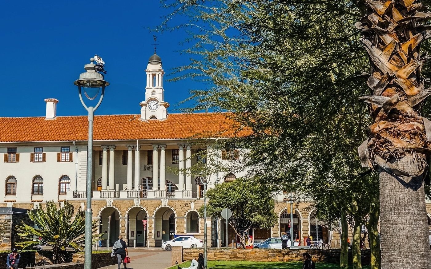 Historic building with a clock tower and arcaded front, beside a lamp post and palm trees.